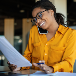 Woman entrepreneur sitting at table with laptop and hand full of papers on phone